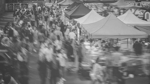 Black and white photograph of a busy outdoor market scene with many people walking past large canopy tents.