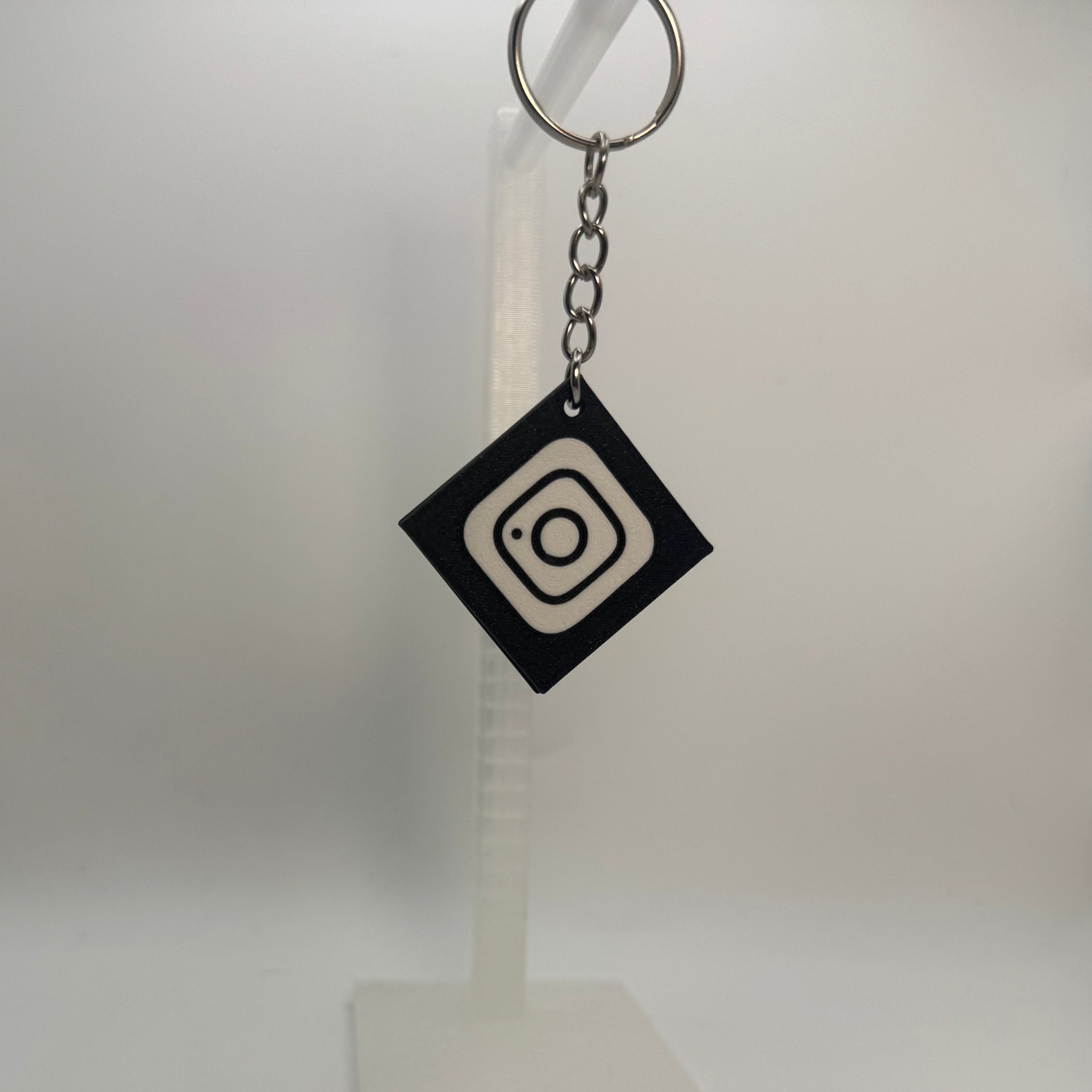 A black diamond-shaped keychain with a white concentric circle logo, hanging from a silver key ring on a clear plastic display stand against a white background.