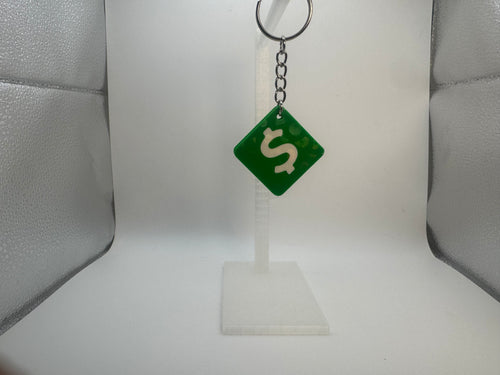 A green diamond-shaped keychain with a white dollar sign, hanging from a silver metal key ring on a clear plastic display stand against a white background.