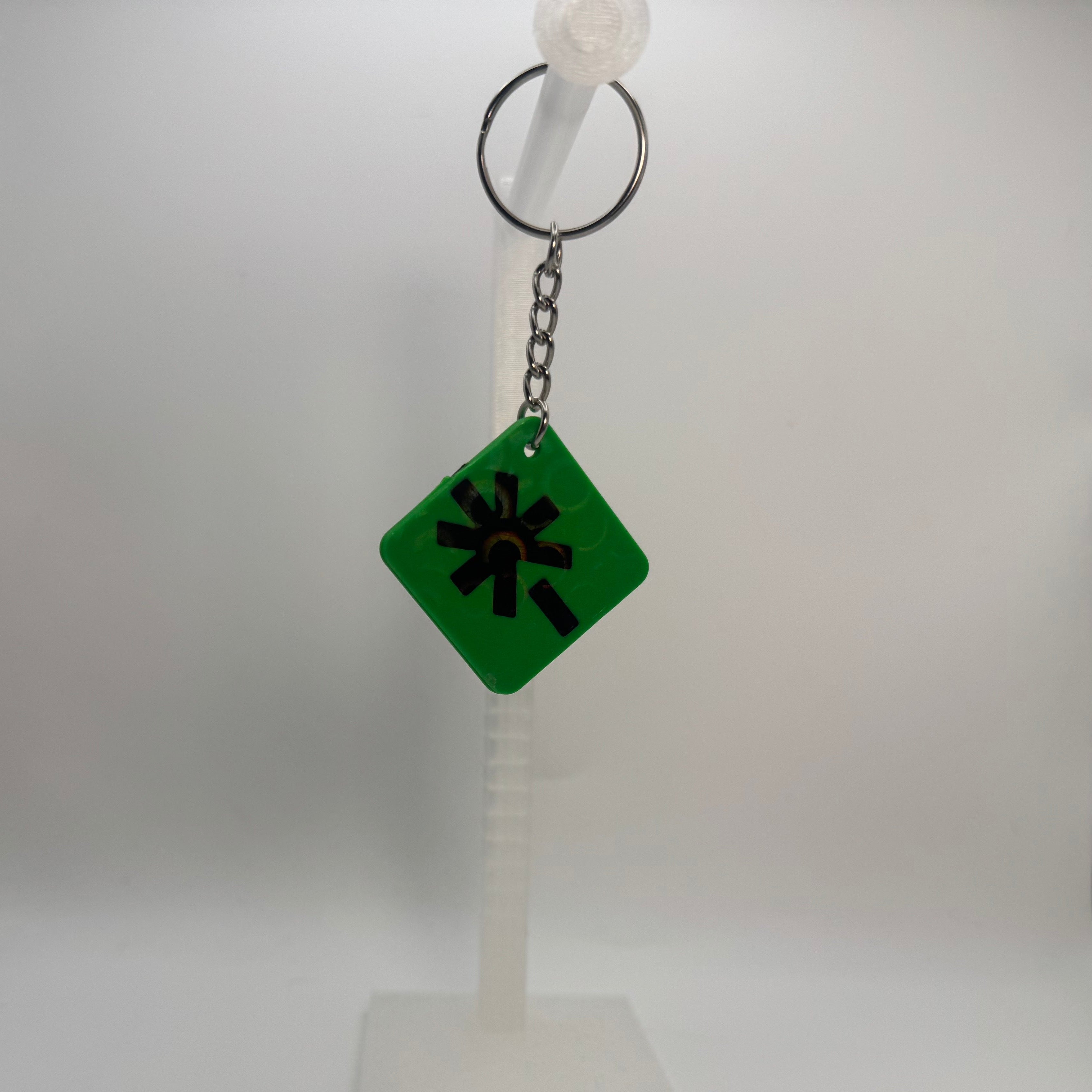 A green diamond-shaped keychain with a black starburst symbol, hanging from a silver metal key ring on a white plastic display stand against a white studio backdrop.