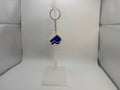 A small white diamond-shaped keychain with a blue logo, hanging from a black ring on a clear plastic display stand against a white studio background.