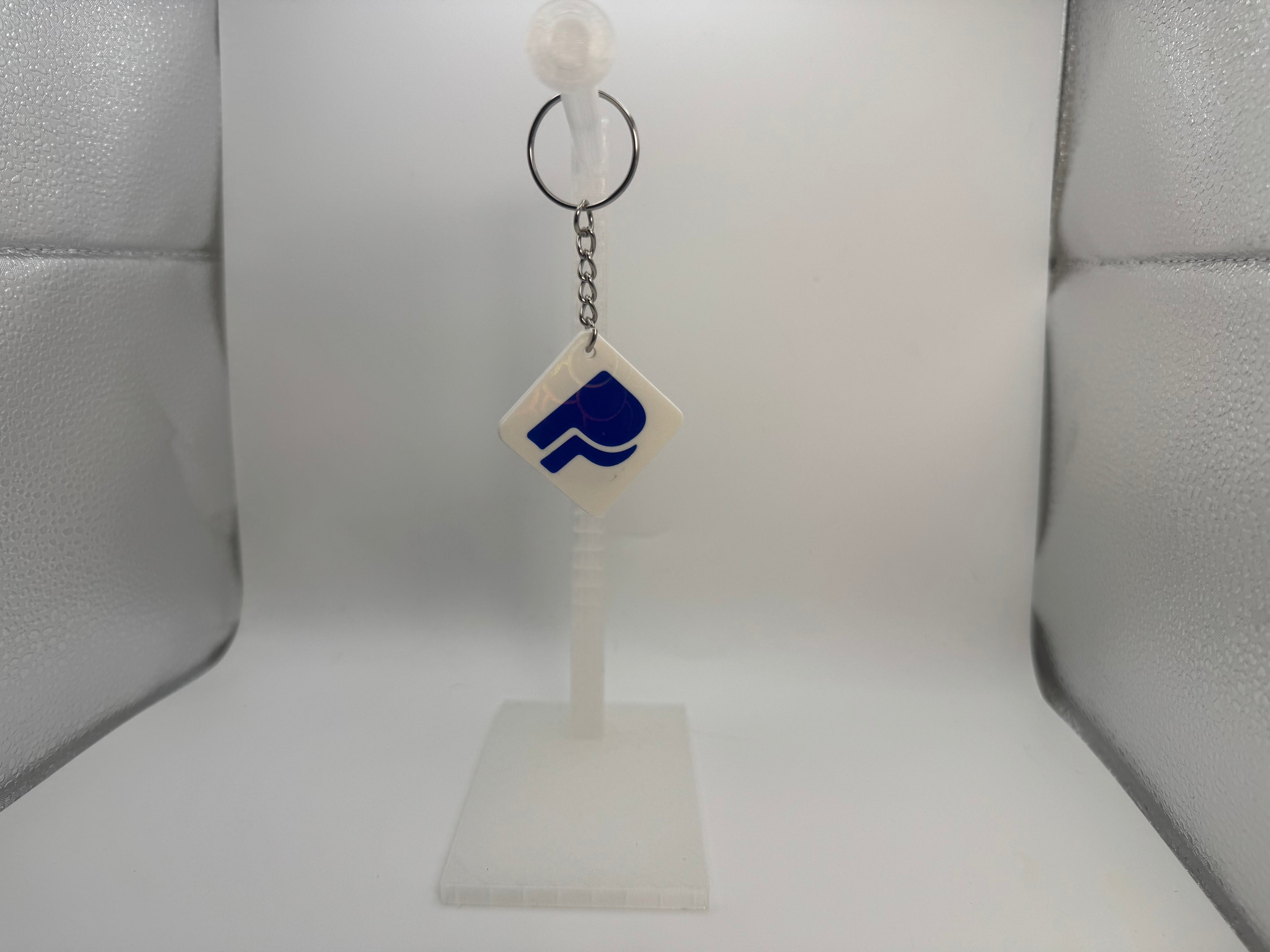 A small white diamond-shaped keychain with a blue logo, hanging from a black ring on a clear plastic display stand against a white studio background.