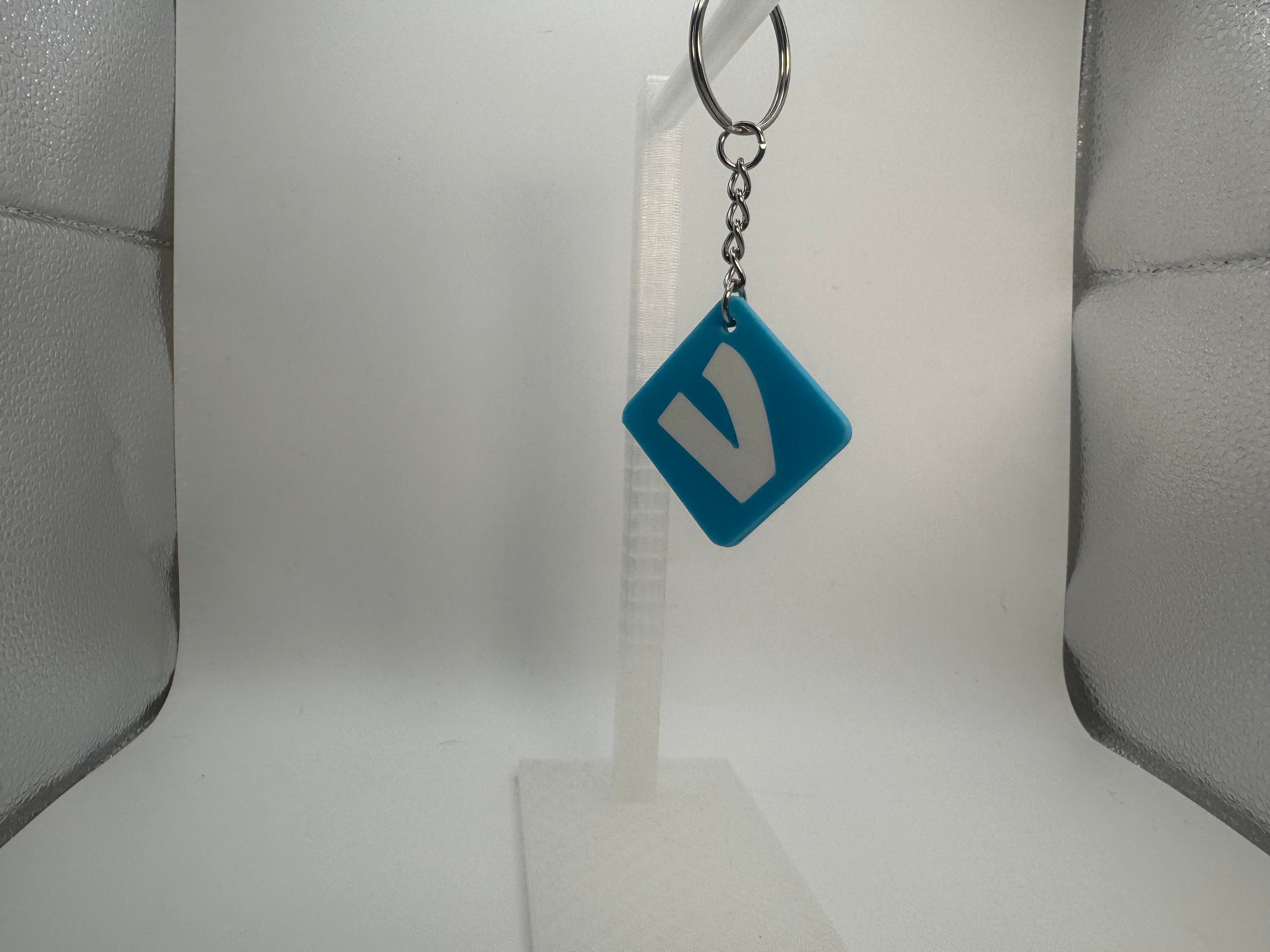 A blue diamond-shaped keychain with a white stylized letter 'V' on a silver split ring, displayed on a clear plastic stand against a white background.